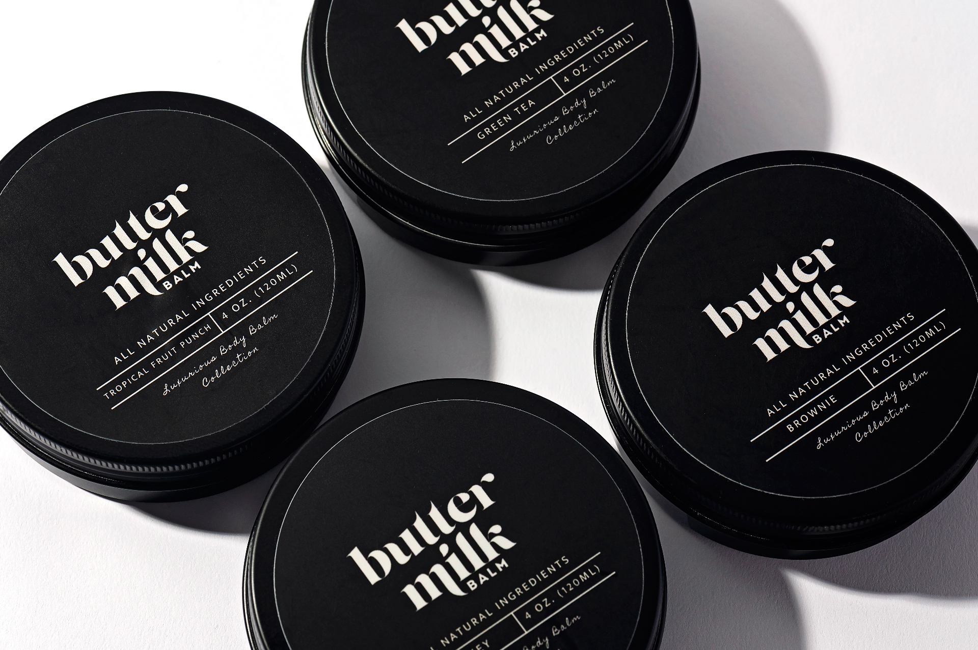 Flatlay of Buttermilk Balm black tins in Tropical Fruit Punch, Green Tea, Brownie, and Whiskey fragrances