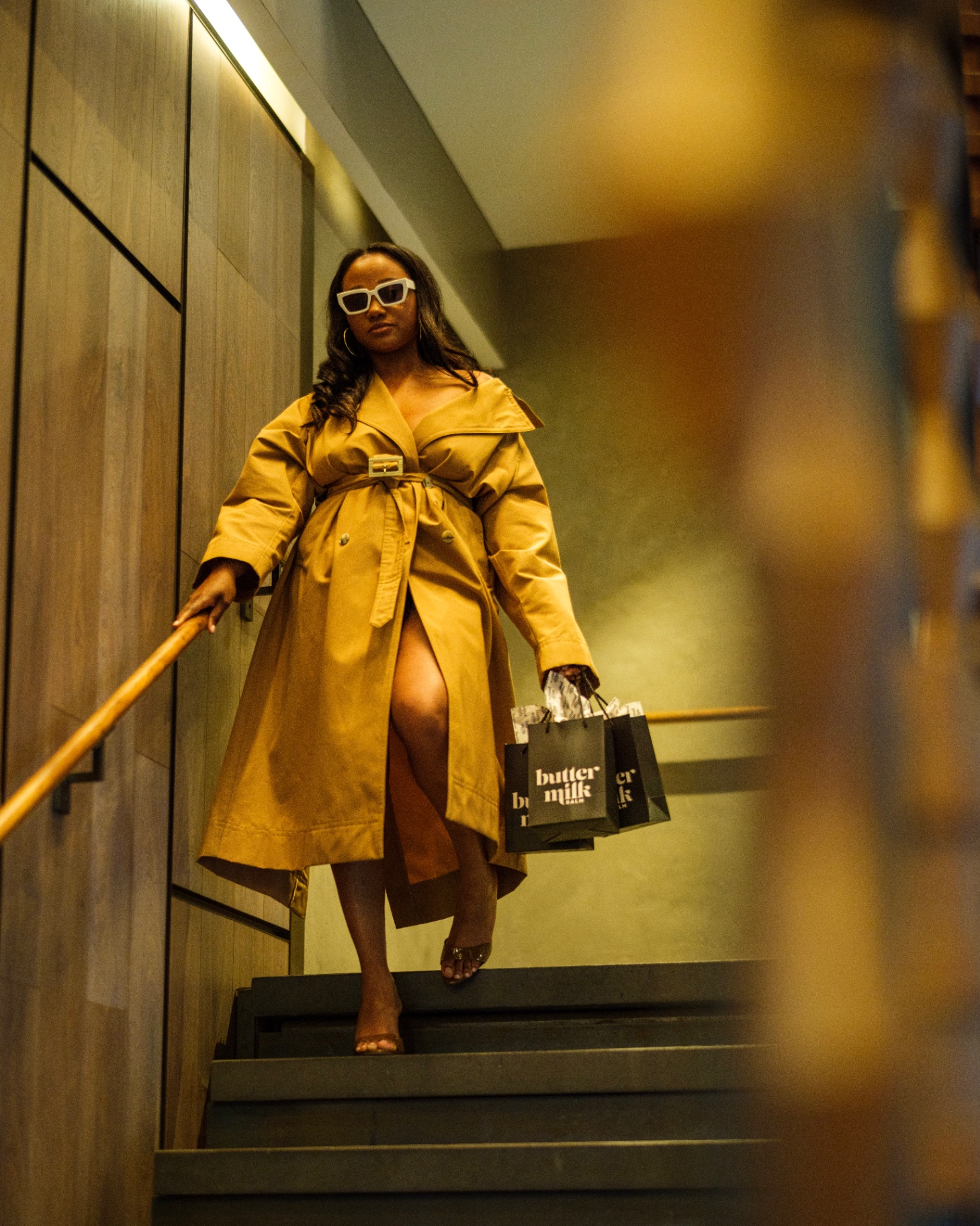 Model in camel trench descending wood-lined stairs with Buttermilk Balm gift bags